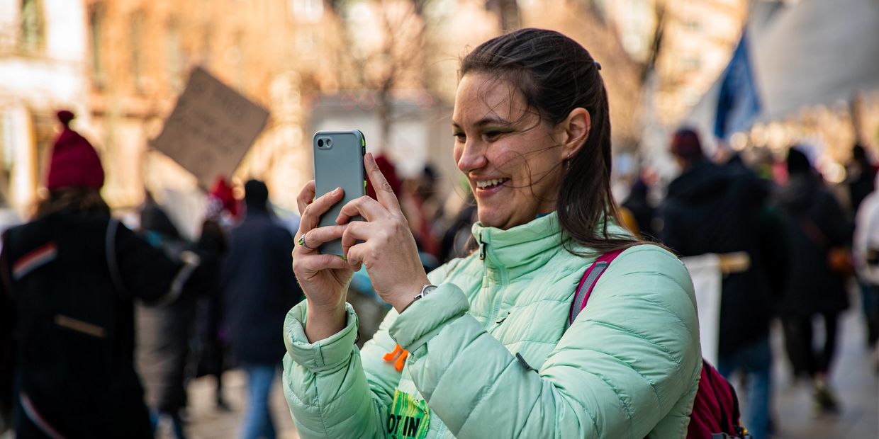 A woman in a green jacket smiling and taking a photo with her phone at an outdoor event.
