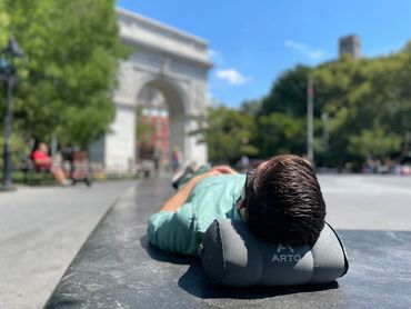 A man laying on a bench with his Arto Pillow - Inflatable in a city park.