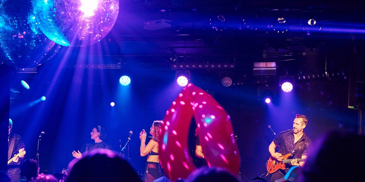 Live band performing under disco lights with audience and inflatable lips.