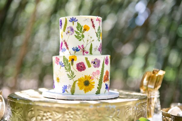 Two-tiered cake decorated with colorful edible flowers on a golden stand.