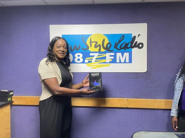 Pastor Patricia stands in front of the New Style Radio banner, holding her book Psalm 91.