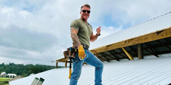 Confident man in work gloves and tool belt standing on a metal roof under a cloudy sky.