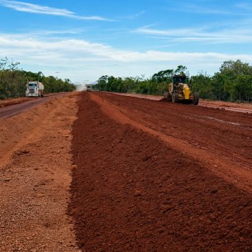 Construction machinery grading red soil on a road under a blue sky.