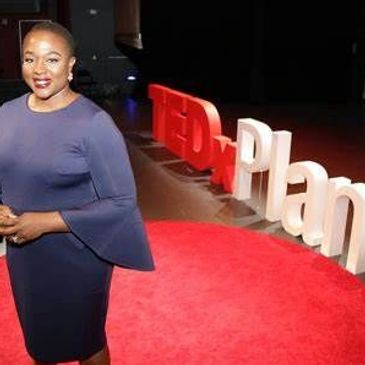 Lola Dada-Olley in a blue dress and short black hair standing on the TEDx Plano stage.