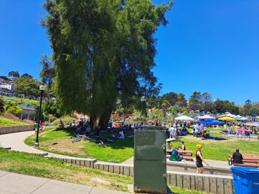 A sunny park day with people relaxing under trees and colorful tents.