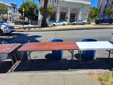 Tables set up on a street with a building and parked cars in the background.