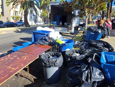 Trash bins and tables filled with garbage bags near a truck on a sunny street.
