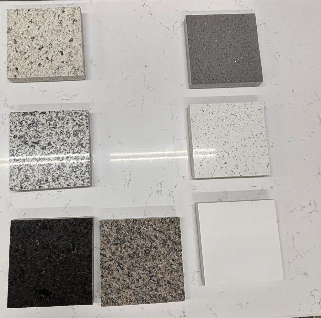 Six square granite and quartz countertop samples on a white marble surface.