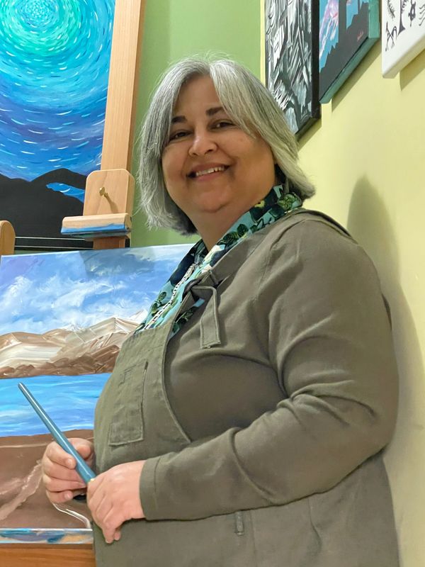 Smiling woman artist holding a paintbrush by her landscape painting.