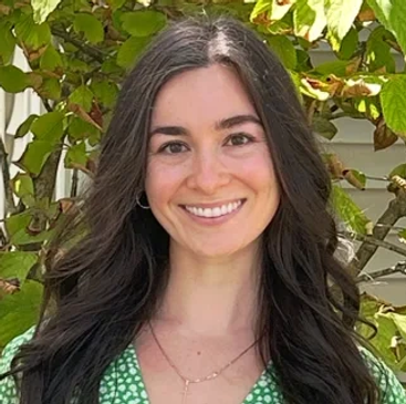 Smiling woman in a green floral dress stands outdoors with lush greenery behind her.