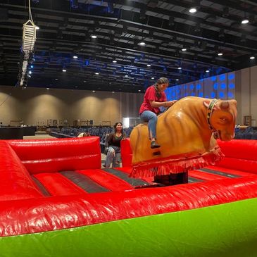 Woman riding a mechanical bull in an indoor arena with red and green inflatable padding.