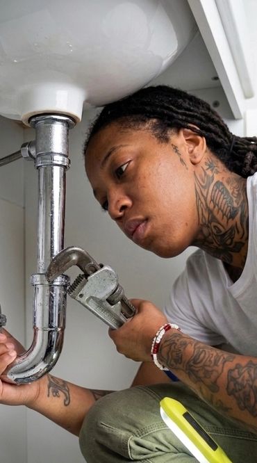 A tattooed person fixing pipes under a sink with a wrench and tools.