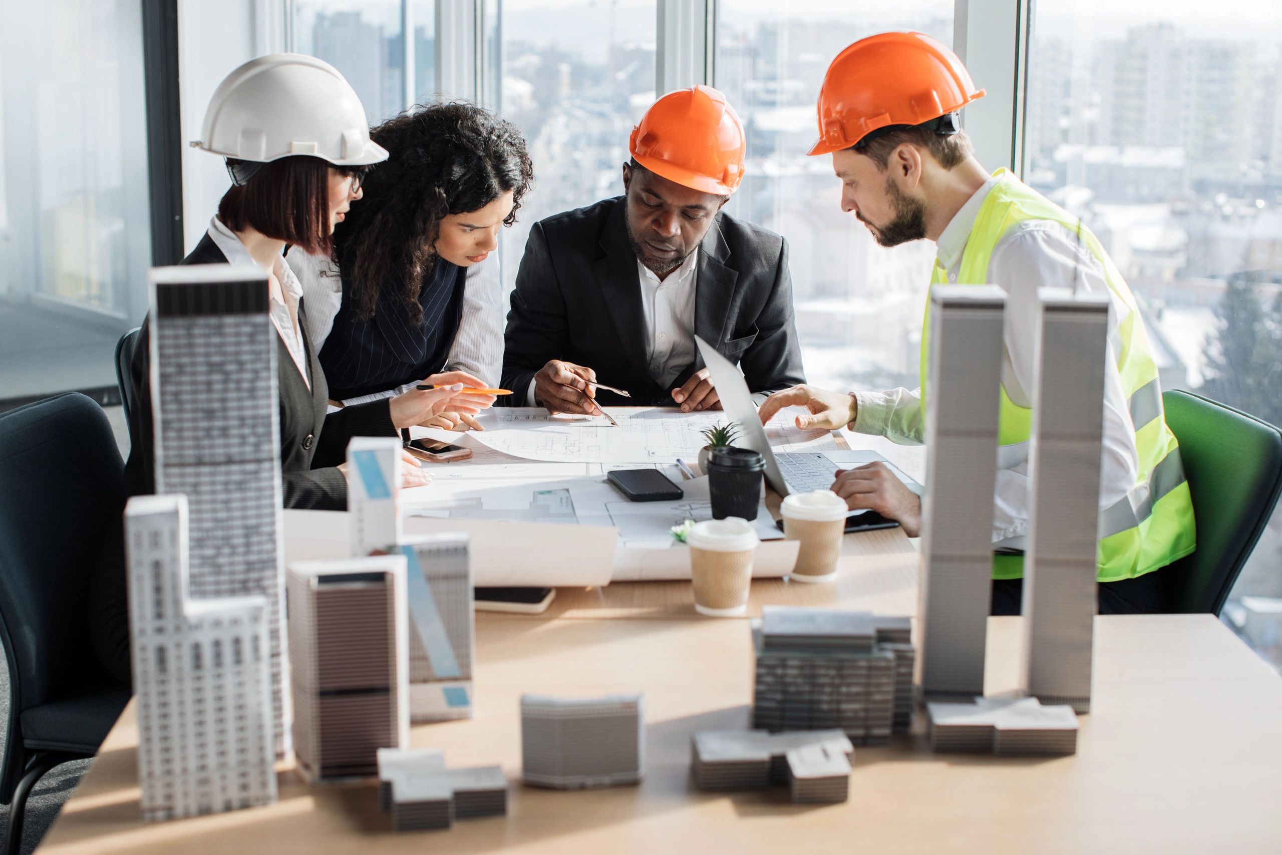 Engineers in hard hats discuss blueprints at a table with building models.