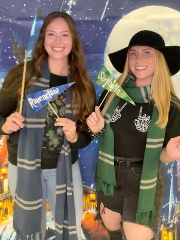 Two women dressed as Ravenclaw and Slytherin from Harry Potter, holding wands and flags.