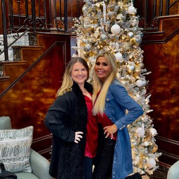 Two women posing happily in front of a decorated Christmas tree in a cozy living room.