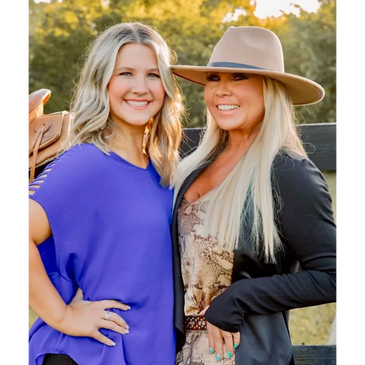 Two smiling women posing outdoors with a saddle and trees in the background.