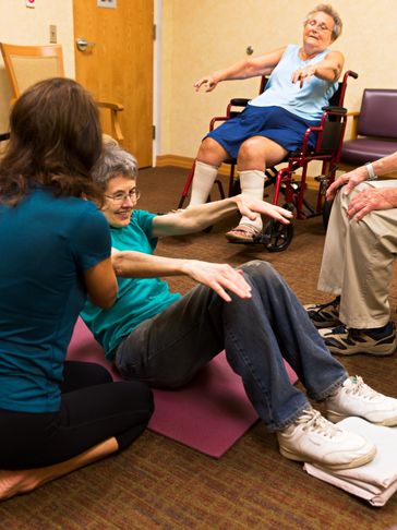 Supporting woman on mat on floor in a core strengthener, with another woman in same pose on a chair.