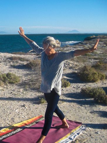 Standing on a yoga mat on the beach with warrior legs and arms outstretched trying to balance.