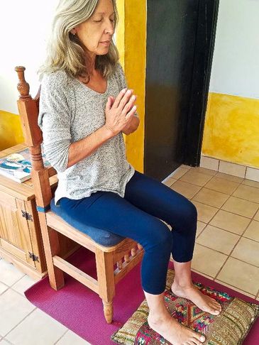 Seated on a wooden hard backed chair with a pillow under both feet, hands in Namaste and eyes closed