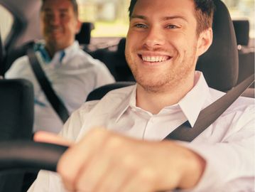 Homem sorridente dirigindo carro com passageiro atrás.
