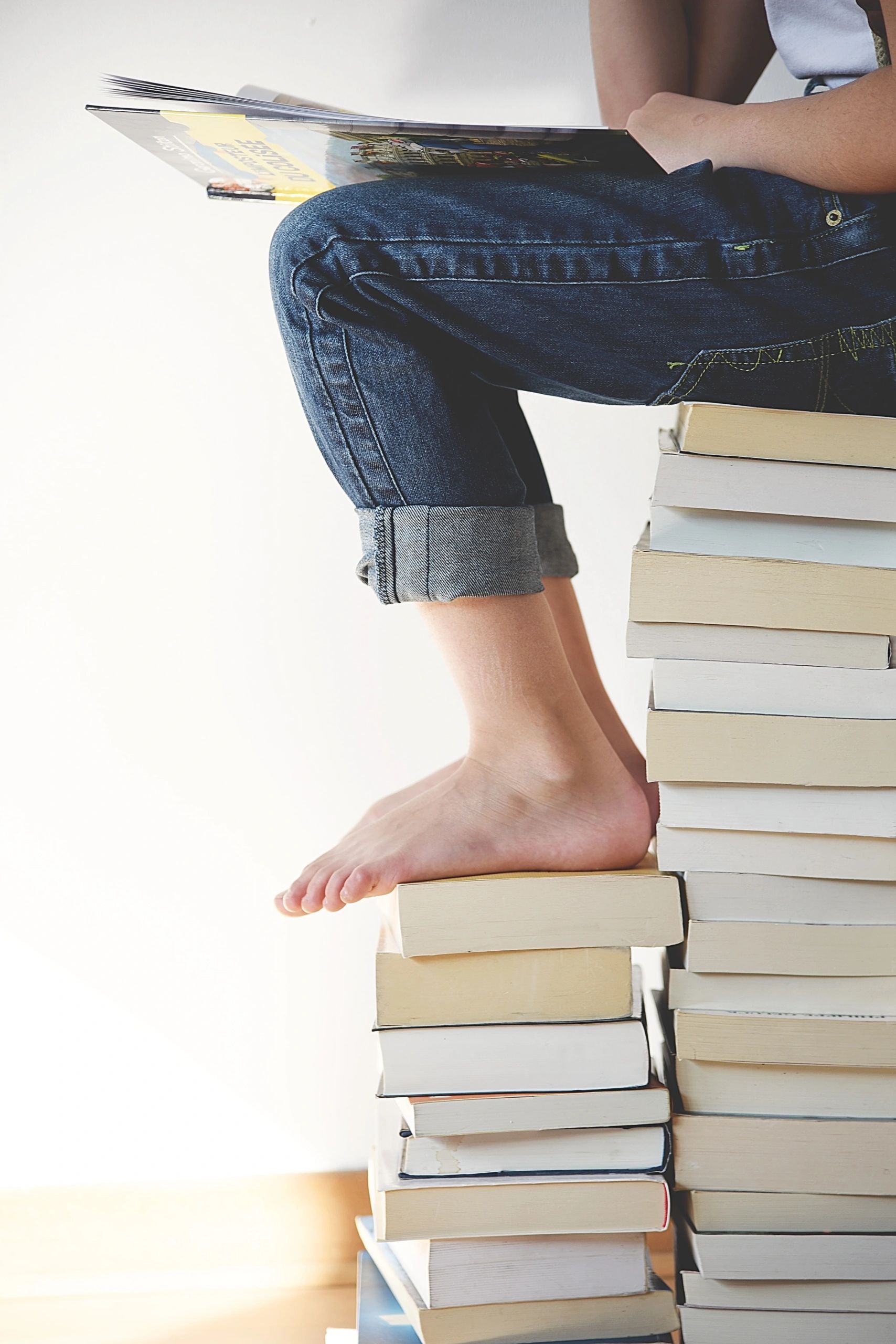 A child is sitting barefoot on top of a pile of books while intently reading a book.