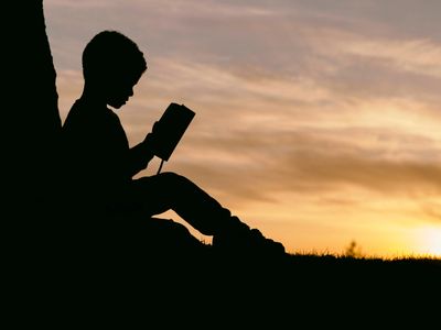 a shadow of a child reading a book while sitting against a wall, at sunset
