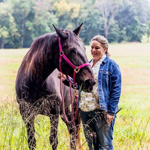 Woman in denim jacket standing with a dark horse in a field.