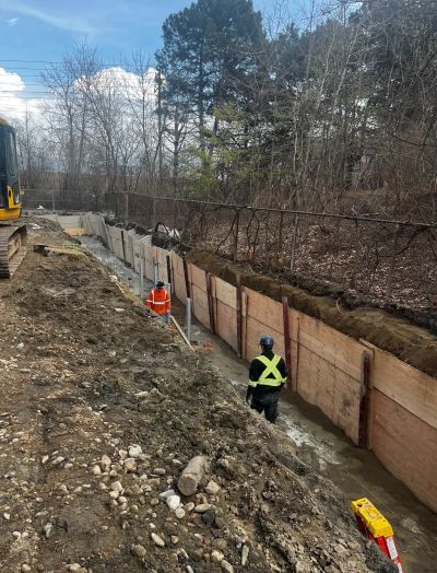 Two construction workers in a trench with wooden supports and machinery nearby.