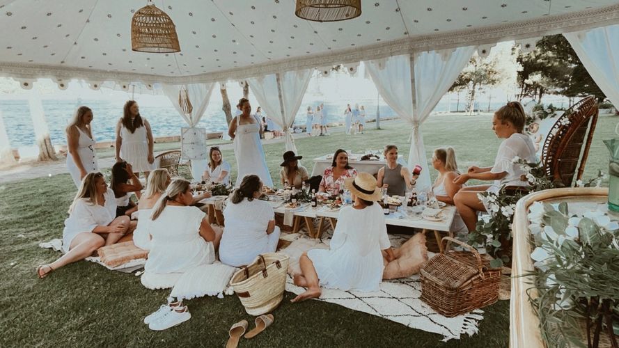 group of women in white outfits sitting at a pretty picnic under a luxury tent