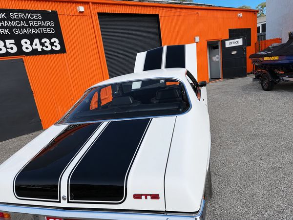 White classic car with black racing stripes at an orange workshop.