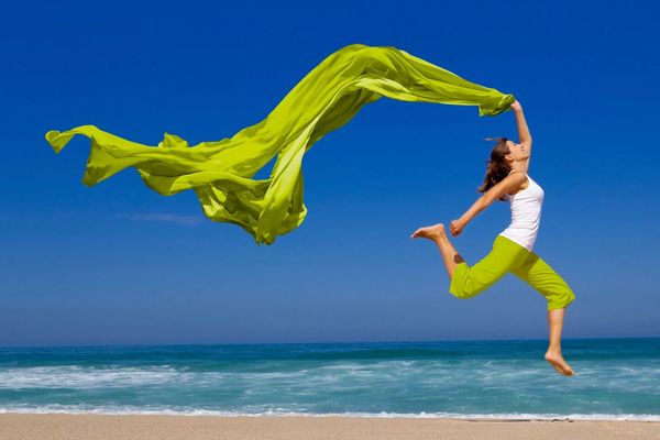 A woman joyfully leaps on a beach holding a flowing green scarf against a clear blue sky.