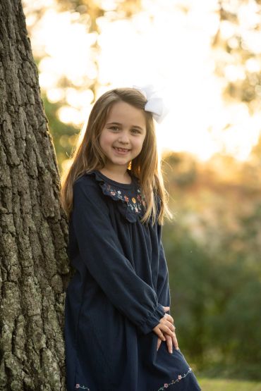 A young girl smiling near a tree with sunlit background.