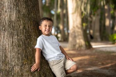 A boy in white t-shirt leaning on a large tree, smiling.
