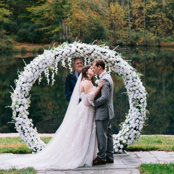 Bride and groom kiss under a white floral arch by a lake during their wedding.