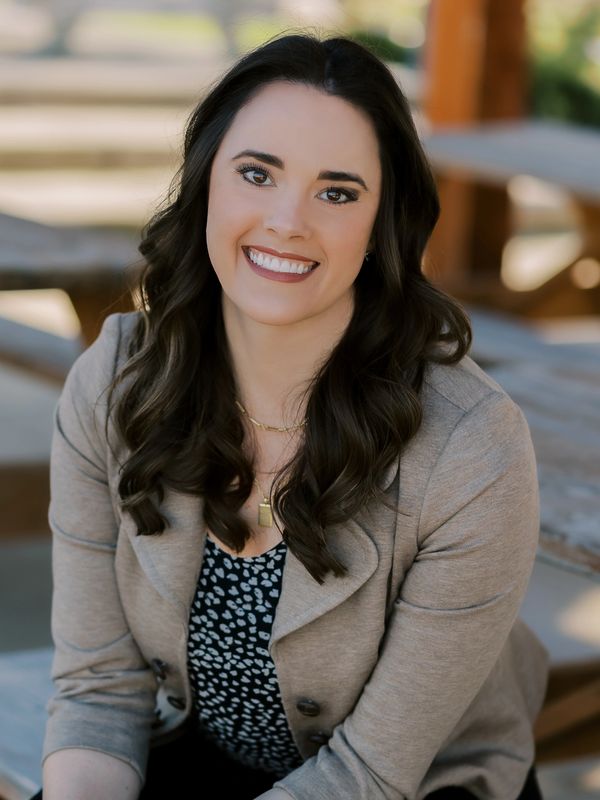 Smiling woman with dark hair wearing a beige jacket and patterned top.