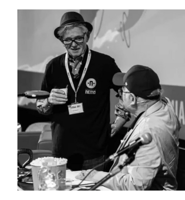 Two elderly men conversing, one holding a drink, in a casual indoor setting.