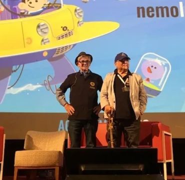 Two men standing on stage with colorful cartoon backdrop.