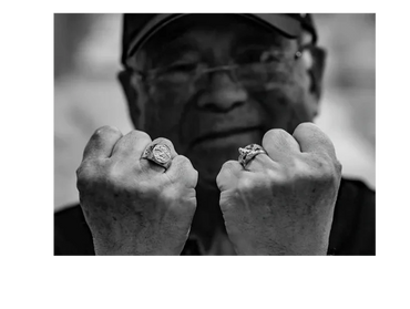 Elderly man shows two large rings on his fists in black and white.