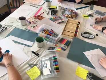 A table scattered with pens, pencils and worksheets with hands colouring various madala patterns