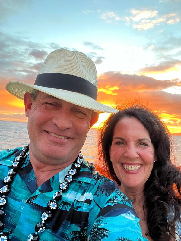 A smiling couple in tropical attire with a sunset ocean background.