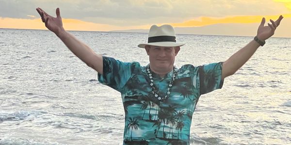 Man in a tropical shirt and hat enjoying the ocean sunset with arms outstretched.