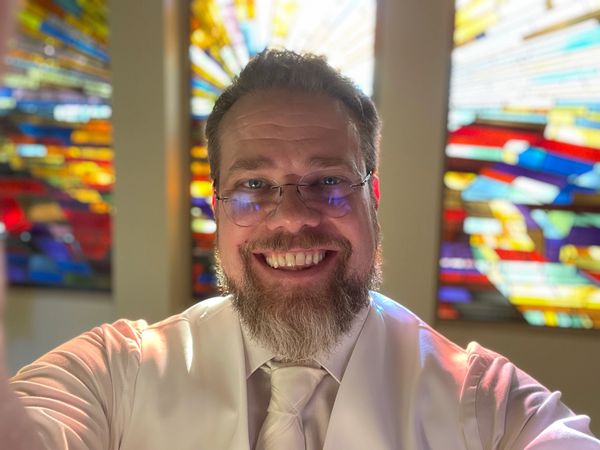 Smiling man with a beard wearing glasses and a white tie in front of stained glass windows.