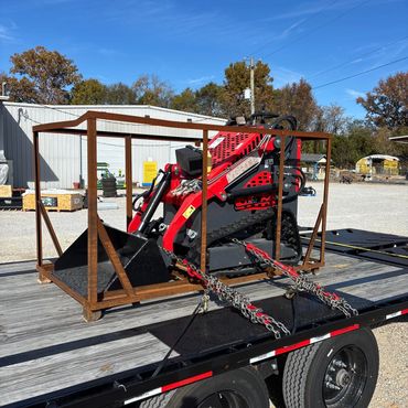 Red compact loader secured on a flatbed trailer with chains.