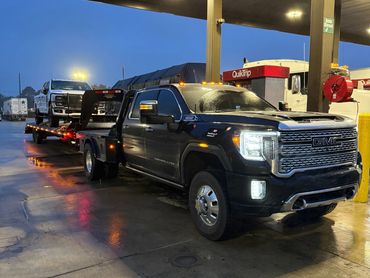 Black GMC Denali HD truck with a flatbed trailer carrying a white pickup at a gas station.