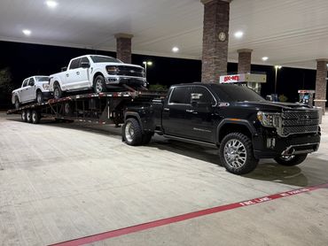 Black GMC truck towing white Ford trucks on a flatbed at night.