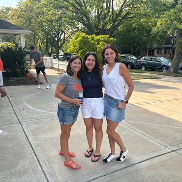 Three women smiling and posing on a driveway with people playing basketball in the background.