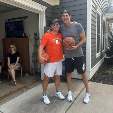 Two men holding basketballs smile outside a house with a woman seated in the background.