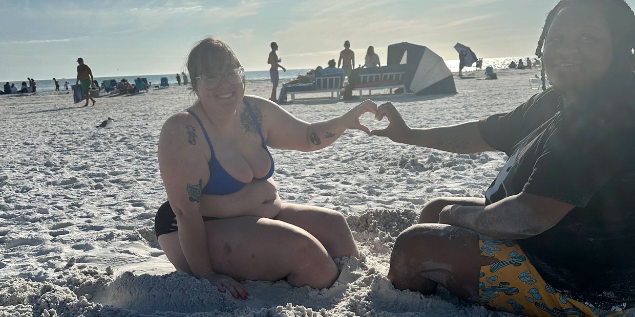 Two people sitting on the sandy beach making a heart shape with their hands.