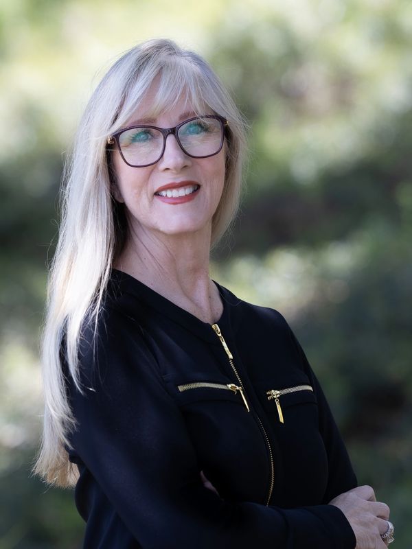 Smiling woman with blonde hair and glasses wearing a black jacket outdoors.