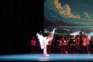 Performer in white doing a balancing pose on stage with musicians.